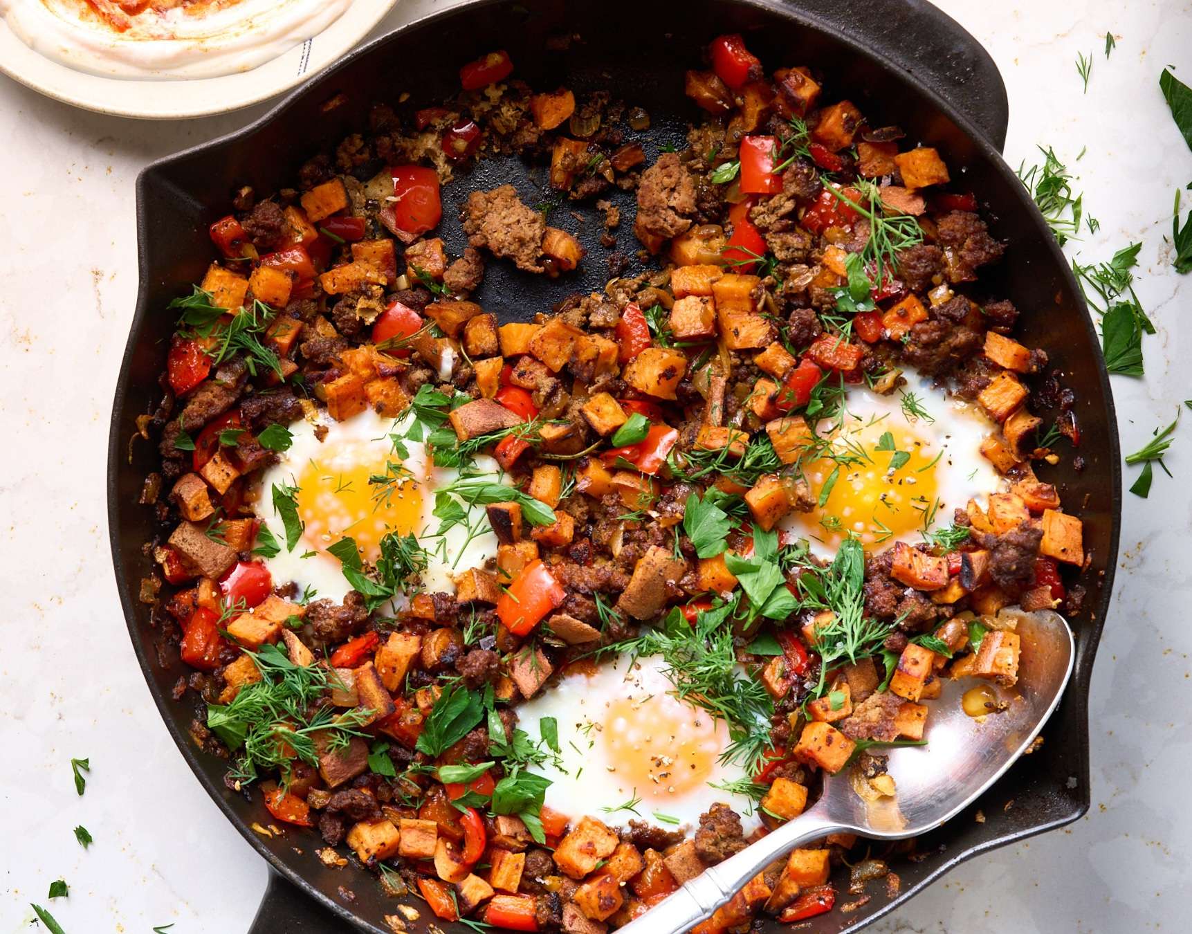 American lamb and sweet potato hash cooked in a skillet with diced red peppers and herbs, topped with sunny eggs and finished with fresh parsley, dill, and a drizzle of harissa yogurt.