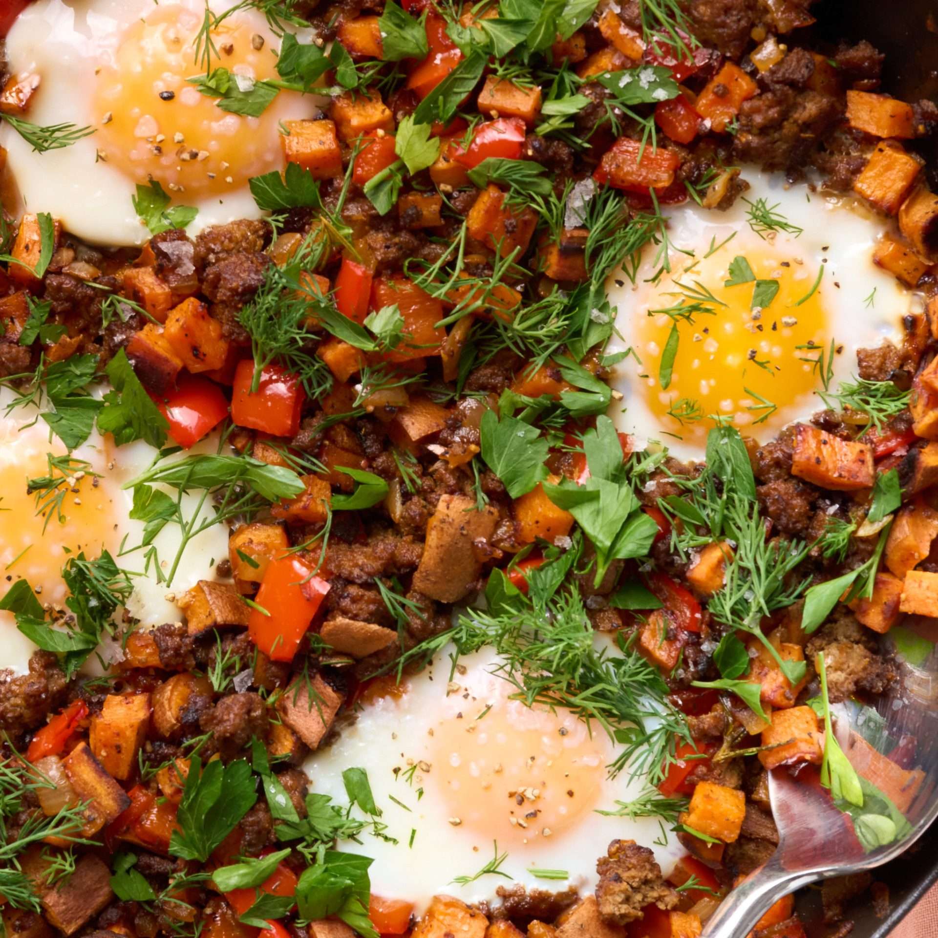 American lamb and sweet potato hash cooked in a skillet with diced red peppers and herbs, topped with sunny eggs and finished with fresh parsley, dill, and a drizzle of harissa yogurt.