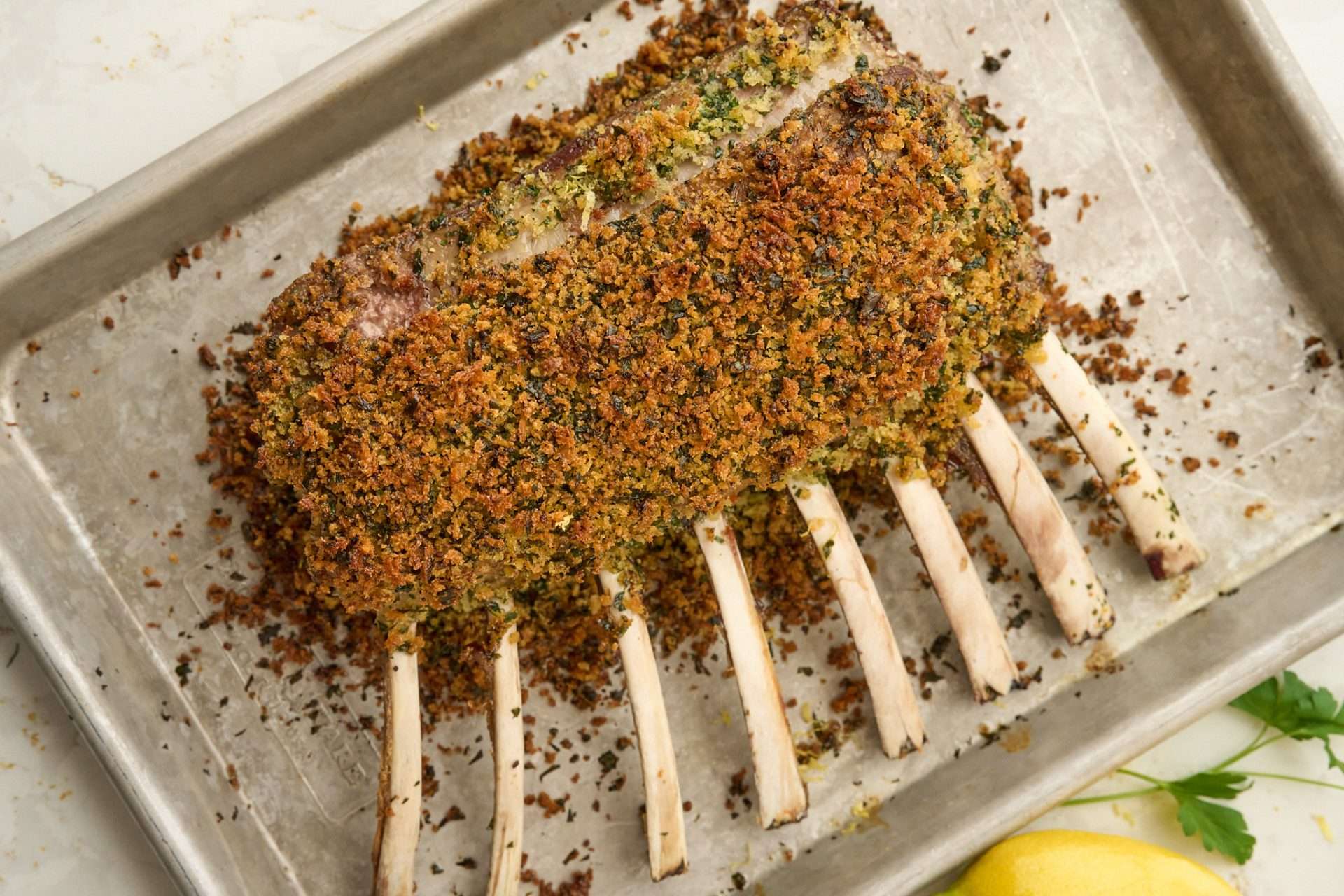 Gremolata crusted rack of American lamb sliced into chops and arranged on a white platter, with a golden herb breadcrumb topping and a juicy pink center.