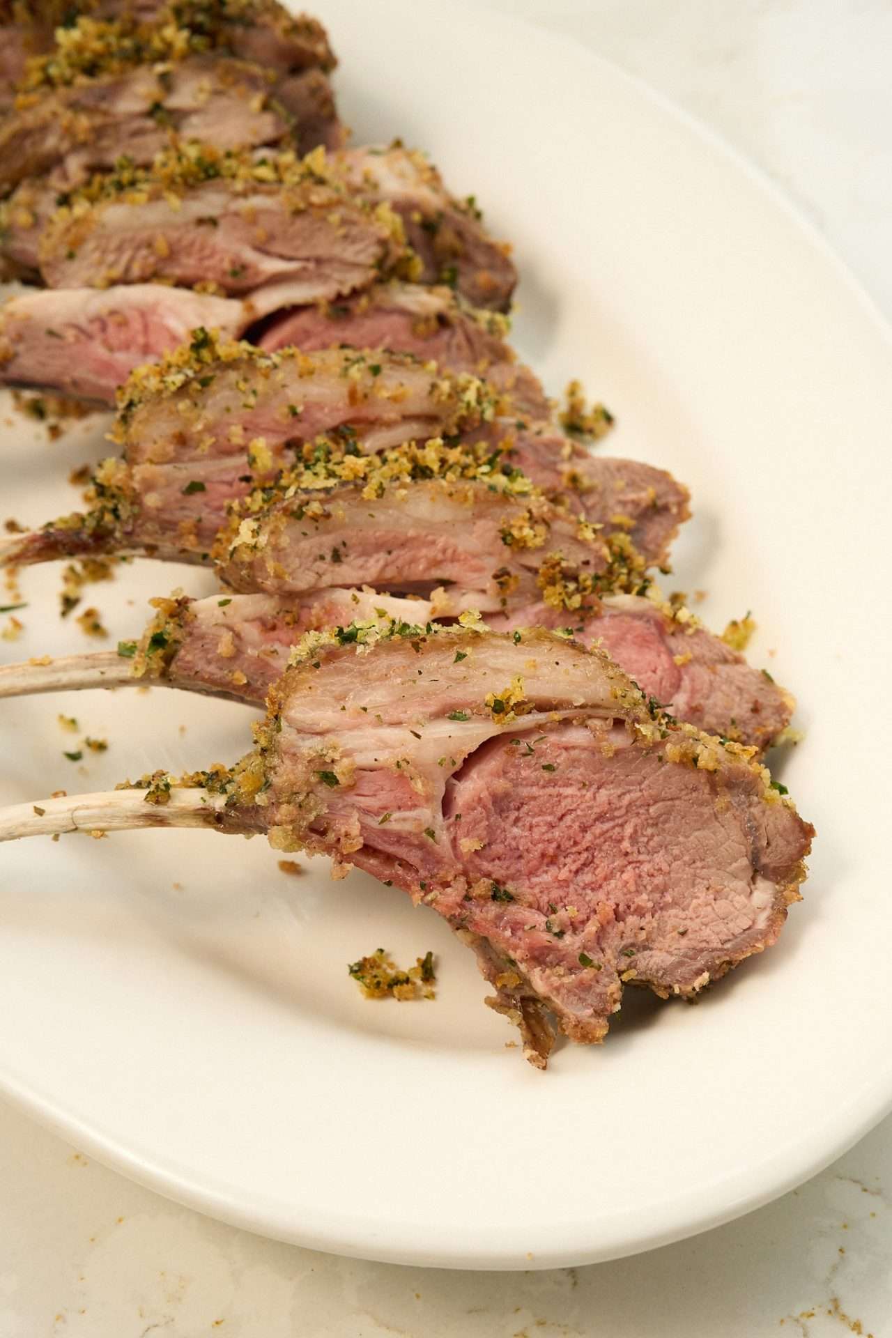 Gremolata crusted rack of American lamb sliced into chops and arranged on a white platter, with a golden herb breadcrumb topping and a juicy pink center.