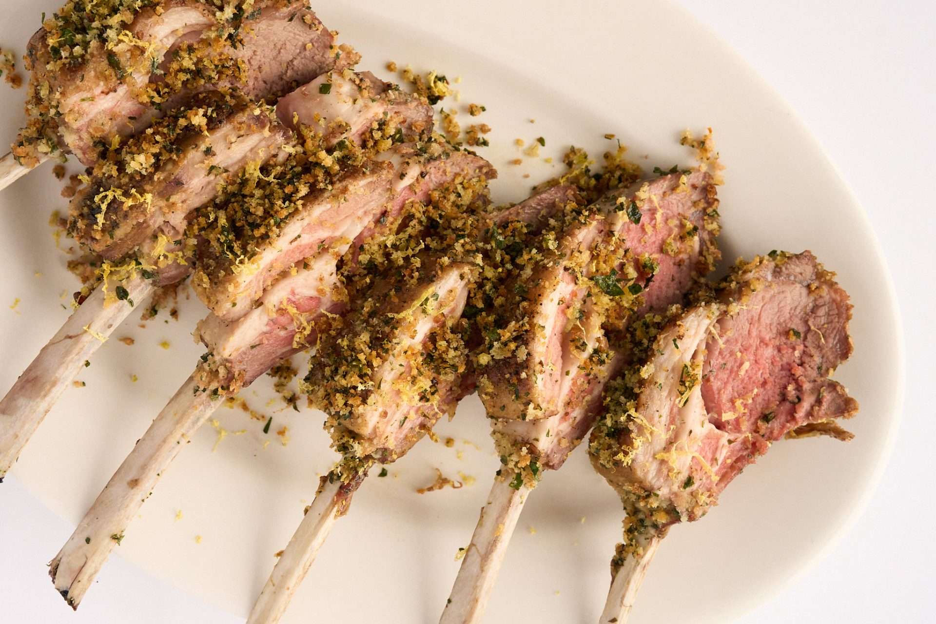 Gremolata crusted rack of American lamb sliced into chops and arranged on a white platter, with a golden herb breadcrumb topping and a juicy pink center.