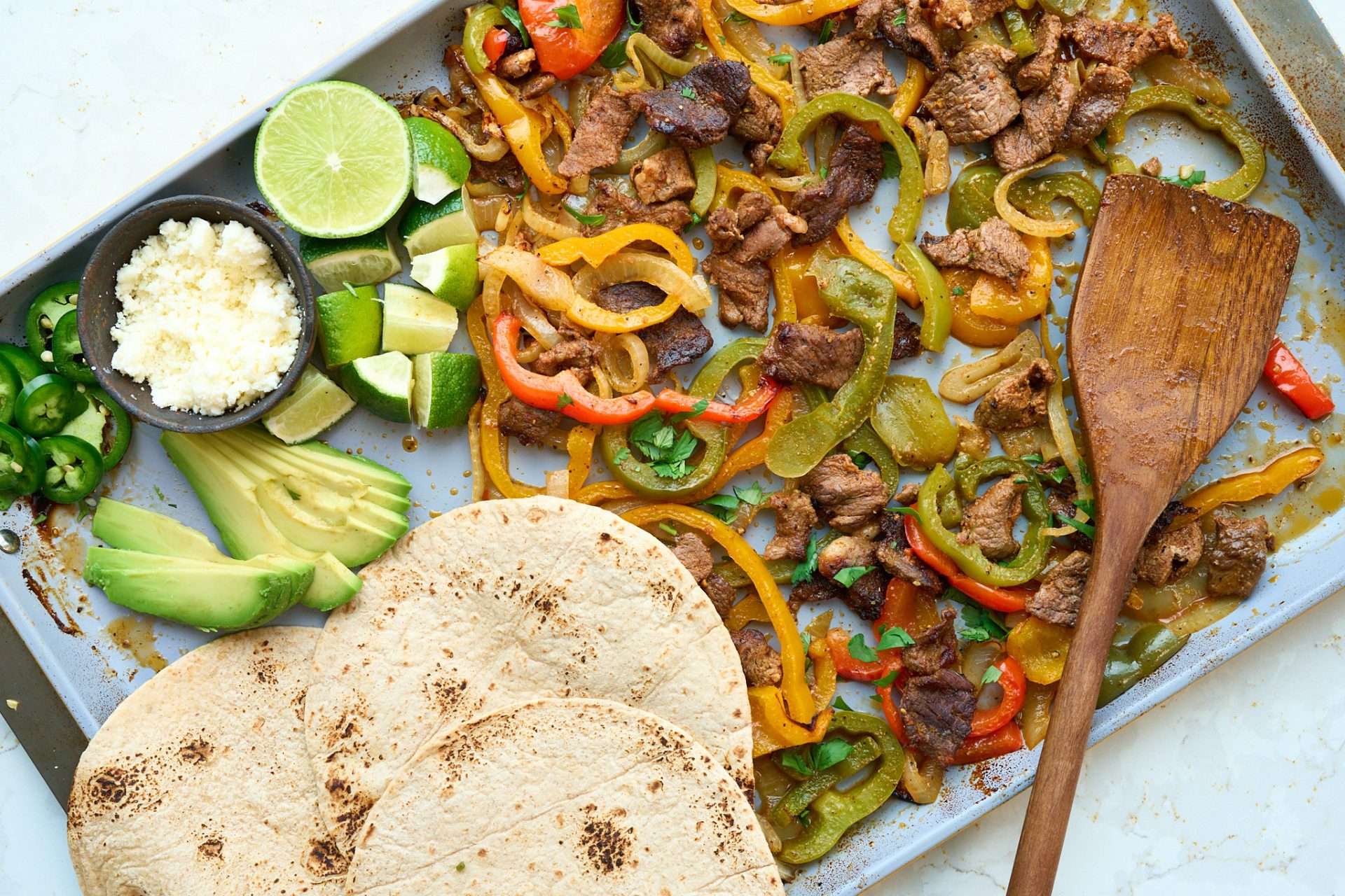 Sheet pan American lamb fajitas served on a warm tortilla with sautéed peppers and onions, sliced lamb, avocado, jalapeños, cilantro, and crumbled cheese, with lime wedges on the side.