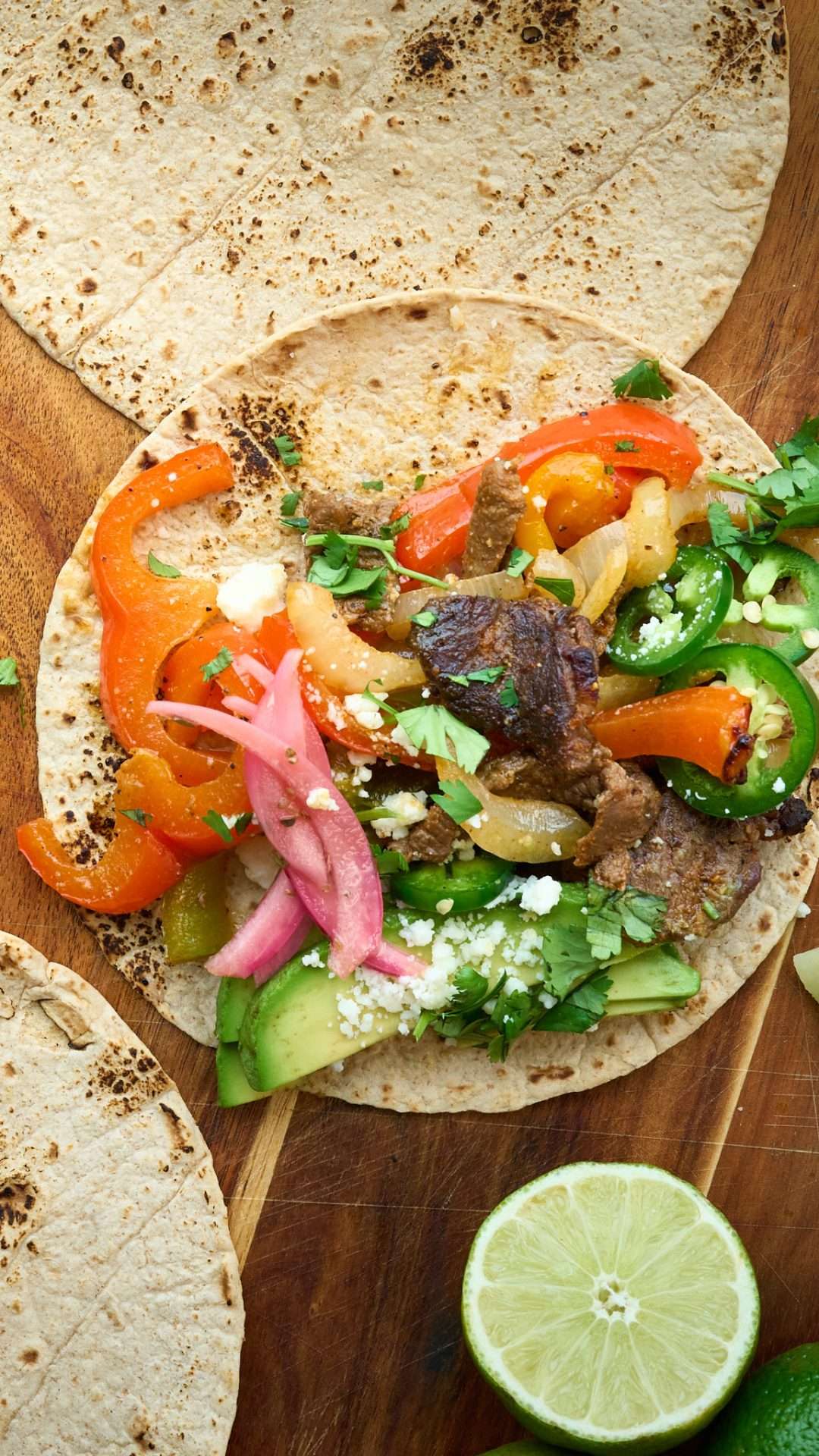 Sheet pan American lamb fajitas served on a warm tortilla with sautéed peppers and onions, sliced lamb, avocado, jalapeños, cilantro, and crumbled cheese, with lime wedges on the side.
