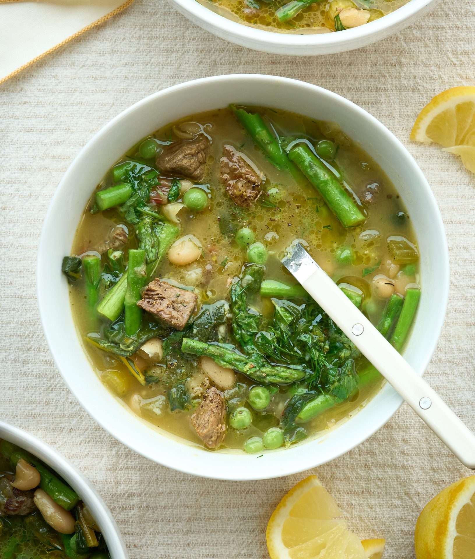 Bowl of lemony American lamb and spring greens soup with tender lamb pieces, asparagus, peas, and leafy greens in a light broth, served with a spoon and a lemon wedge on the side.