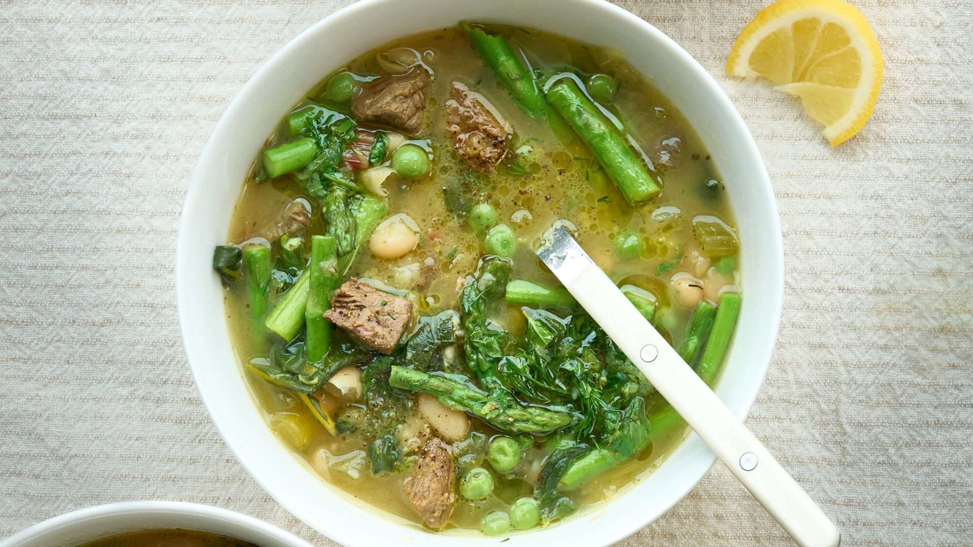 Bowl of lemony American lamb and spring greens soup with tender lamb pieces, asparagus, peas, and leafy greens in a light broth, served with a spoon and a lemon wedge on the side.